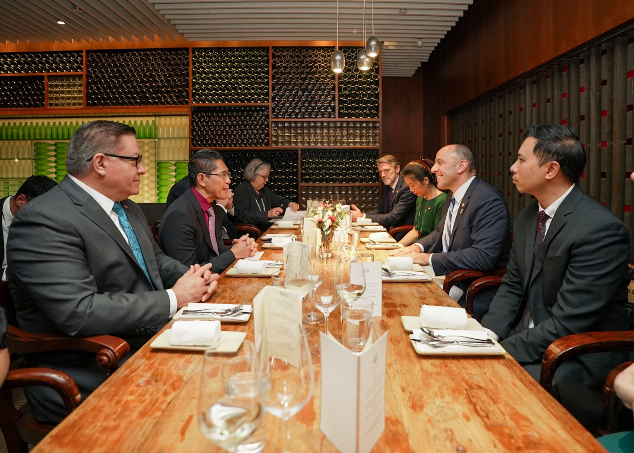 People in suits at long wooden table with wine bottles on wall.